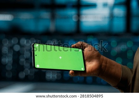 A software developer checks server performance on a mockup smartphone and monitors system alerts through a green screen display.