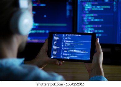 Software developer freelancer with white headphones and glasses working with program code: C++, Java, Javascript on wide displays and tablet at night. Develops new web, desktop, mobile application.