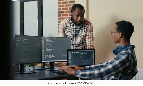 Software Developer Sitting At Desk With Multiple Screens And Laptop Running Code Talking With Colleague About Artificial Intelligence Algorithm. Software Developers Doing Teamwork In It Office.