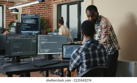 Software developer writing code on laptop and computer keyboard looking at multiple screens with programming language is interrupted by coworker asking for advice. Cloud programers doing teamwork.