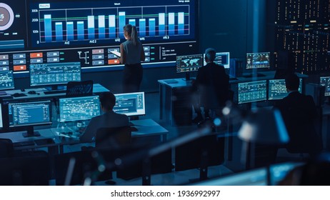 Software Engineers Working In A Room With A Big Screen In A Modern Monitoring Office With Live Analysis Feed With Charts. Monitoring Room Big Data Scientists And Managers Sit In Front Of Computers.