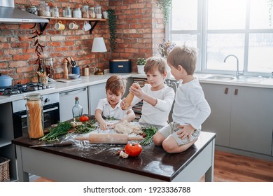 Soiled And Young Kids Dressed In Casual Clothing Learning To Cooking And Play With Flour In Modern Kitchen In Sunny Day.