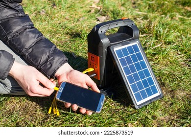 A solar panel charges a phone on a nice sunny day. The phone is in the hands of a man who is charging it. Portable powerbank for hiking and tourism. - Shutterstock ID 2225873519