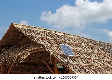 A solar panel on the roof of a thatched hut against blue sky and clouds.