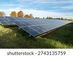 Solar panels array in field under blue sky with scattered clouds, groundmounted racks on grassy meadow, autumn trees along horizon, angled panels reflecting sunlight, commercial