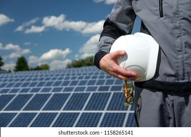 Solar Panels Engineer Holding White Protective Cask And Glasses, Hand Close, Solar Pannel Field Background. Hot Sunny Summer Weather. Copy Space. Concept Of Renewable And Clean Energy, Technology.