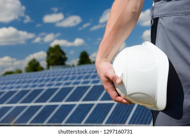 Solar Panels Engineer Holding White Protective Cask, Hand Close, Solar Pannel Field And Sky Background. Hot Sunny Summer Weather. Copy Space. Concept Of Renewable And Clean Energy, Technology.