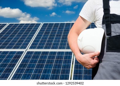 Solar Panels Engineer Wearing Grey Outfit Holding White Protective Cask, Hand Close, Solar Pannel Field And Sky Background. Copy Space. Concept Of Renewable And Clean Energy, Technology.