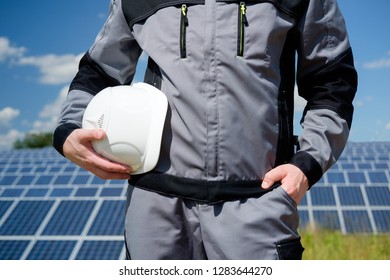 Solar Panels Engineer Or Worker In Grey Outfit Holding White Protective Cask, Hand Close, Solar Pannel Field And Sky Background. Hot Summer Weather. Concept Of Renewable And Clean Energy, Technology.