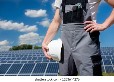 Solar Panels Engineer Or Worker In Grey Outfit Holding White Protective Cask, Hand Close, Solar Pannel Field And Sky Background. Copy Space. Concept Of Renewable And Clean Energy, Technology.