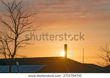 Solar panels installed on a rooftop with a chimney and leafless trees silhouetted against a warm orange sunset sky with soft clouds.