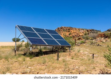 Solar Panels Mounted On A Metal Frame In An Isolated Semi Arid Area. Rocky Cliffs Behind The Panels. Clear Blue Sky 