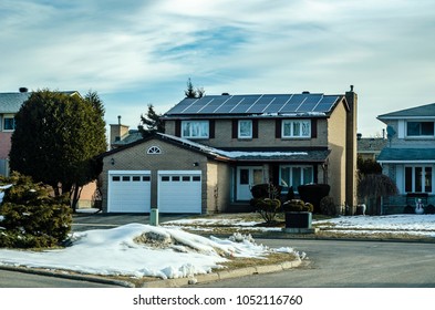 Solar Panels On A Brown Brick Suburban House Rooftop And Garage With Snow On The Ground And Cloudy Blue Sky Background