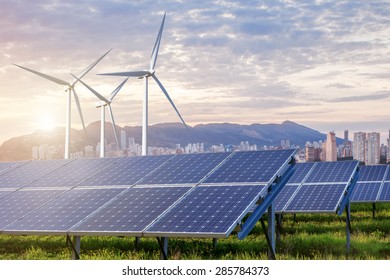 Solar Panels And Wind Turbines Under Sky And Clouds With City On Horizon. Sunrise