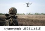 A soldier in camouflage observes a drone hovering above an open field. The drone represents modern warfare technology, while the soldier maintains a vigilant stance.