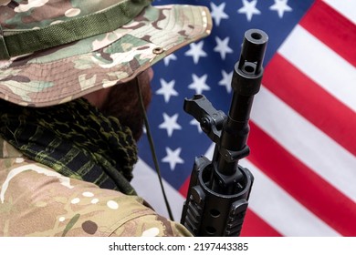 A Soldier In Camouflage Uniform And Panama Hat With An Automatic Rifle On His Shoulder Against The Background Of The American Flag., Concept: Military Operations, Soldier Of Fortune, US Army.