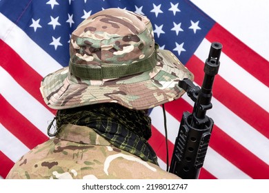 A Soldier In Camouflage Uniform And Panama Hat With An Automatic Rifle On His Shoulder Against The Background Of The American Flag., Concept: Military Operations, Soldier Of Fortune, US Army.