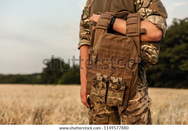 Soldier man standing against a field. Soldier\
in military outfit with bulletproof vest. Photo of a soldier in\
military outfit holding a gun and bulletproof vest on orange desert\
background.