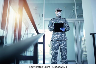 Soldier In Military Camouflage Uniform With Rubber Gloves And Mask Guarding In Front Of Hospital's Door And Controlling Who Gets In And Out. Military Helping In Fight Against Coronavirus Or COVID-19.