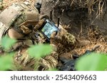 A soldier in tactical gear operates a control tablet in the field, using technology for reconnaissance with an aerial view display on the screen.