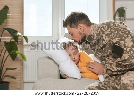 Soldier in Ukrainian military uniform kissing his daughter while she sleeping on sofa at home. Family reunion