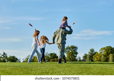 Soldier Is Walking With His Family In The Grass, Back View. Patriotic Family Waving Flags Outdoors On The Park Meadow Grass.