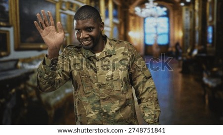 Soldier waves hand and smiles inside an ornate building hall wearing camouflage uniform and raised palm gesture; pride duty welcome.