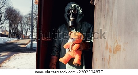Soldier wearing anti nuclear clothing holds toy bear in hand and looking from open metal door of abandoned building