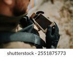 Soldier wearing tactical gloves is using a rugged mobile phone attached to his vest during a military operation, highlighting the integration of technology in modern warfare