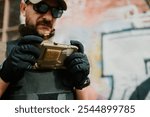 Soldier wearing tactical gloves and vest, uses a ruggedized mobile phone in an urban setting with graffiti in the background, showcasing the integration of technology in modern warfare