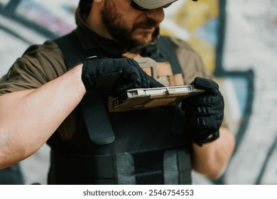 Soldier wearing a tactical vest and gloves uses a ruggedized tablet computer in an urban setting, potentially coordinating operations or accessing critical information - Powered by Shutterstock