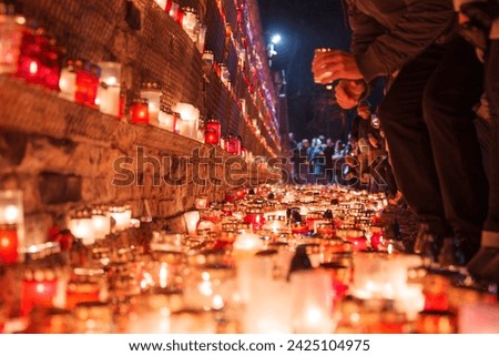 A solemn gathering in Latvia, with red candle holders illuminating the scene, signifies a respectful commemoration of Independence Day, as people in warm attire add to the glowing tribute.
