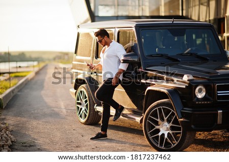 Solid asian man in white shirt and sunglasses posed near black mafia suv car with mobile phone.