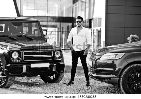 Solid asian man in white shirt and sunglasses posed\
near black mafia suv\
cars.