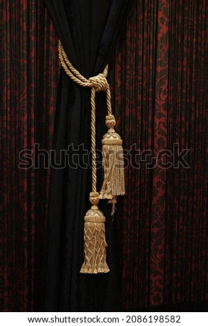 A solid black drape wrapped with a gold cord adorned with a gold tassel against the backdrop of a patterned black and red drape.