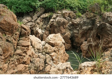 The solid brownish rock cliffs with cracked textures contrast with the lush green trees and bushes above, a beautiful geological natural landscape.