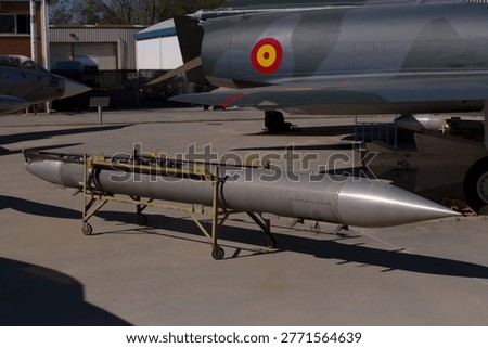 A solid rocket is placed on a stand at a military airbase. Fighter jets are parked in the background, showing a mix of colors and shapes under bright daylight.