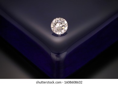 A solitaire, faceted, clear, round diamond sits on a reflective dark blue glass block background.