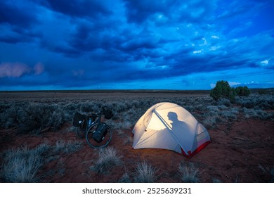 Solitary camping under a moody sky in the remote wilderness. A tent illuminated by warm light, with a bicycle nearby, creating the perfect adventure setup for an unforgettable night outdoors - Powered by Shutterstock