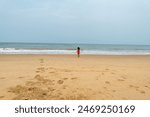 A solitary child in red shorts walks along the shoreline, leaving footprints on the vast sandy beach under an overcast sky
