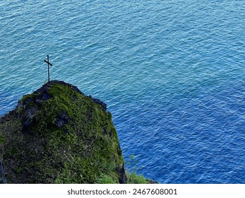 Solitary Cross on Moss-Covered Cliff Overlooking Calm Blue Ocean Waters - Powered by Shutterstock
