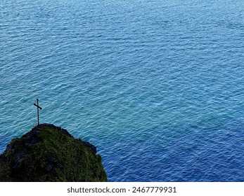 Solitary Cross on Moss-Covered Cliff Overlooking Calm Blue Ocean Waters - Powered by Shutterstock