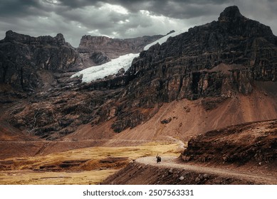 A solitary cyclist rides along a rugged, dirt road that winds through a dramatic and foreboding mountain landscape in the Peruvian Andes. The towering cliffs and jagged peaks loom large. - Powered by Shutterstock