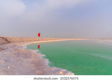 A solitary figure in red stands on a tranquil salt bank, reflecting in calm, emerald waters under a hazy sky. - Powered by Shutterstock