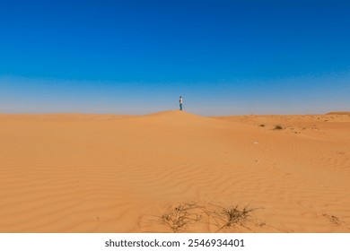 A solitary figure stands atop a sand dune in the Wahiba Desert during midday surrounded by expansive golden sands and clear blue skies - Powered by Shutterstock