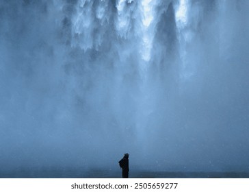 A solitary figure stands in awe at the base of a powerful waterfall, enveloped in mist and the overwhelming force of nature, capturing the scale and majesty of the natural world. - Powered by Shutterstock