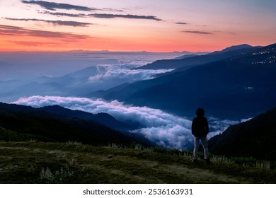 A solitary figure stands on a mountain ridge, mesmerized by a stunning sunrise. The sky glows with orange and gold, while a tranquil lake of clouds fills the valleys, evoking peace and wonder - Powered by Shutterstock