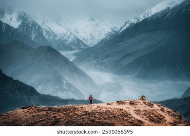 A solitary figure stands on a rugged hilltop against a backdrop of towering, mist-covered mountains. The dramatic contrast between the person and the vast, snowy peaks emphasizes the scale and majesty - Powered by Shutterstock