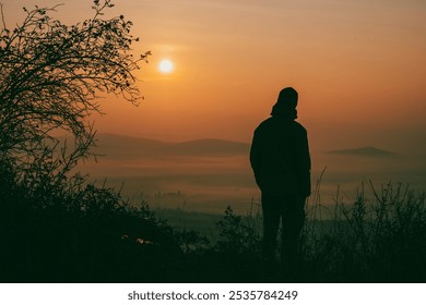 A solitary figure stands softly silhouetted against the warm, glowing hues of an autumn sunrise. Mist rises from the valley as nature awakens to a new day filled with tranquility. - Powered by Shutterstock