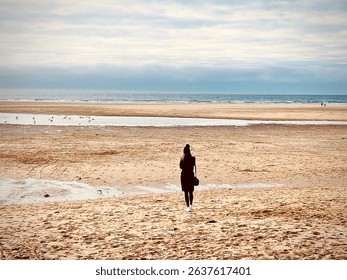 A solitary figure walks toward the sea across a wide expanse of sandy beach under a soft, cloud-dappled sky. The ocean tide is low, revealing a vast stretch of wet and dry sand. - Powered by Shutterstock - Get 15% off with code: PIKWIZARD15